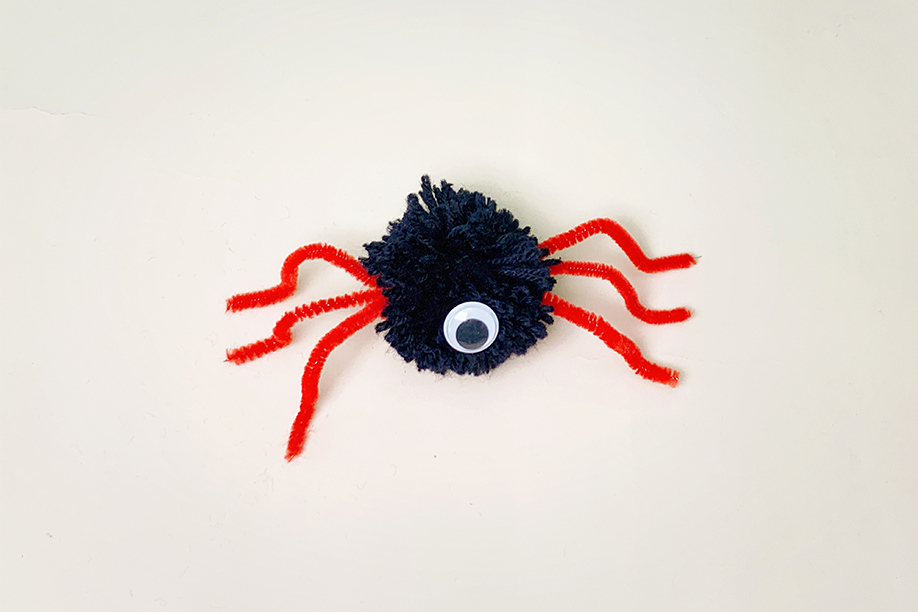 A completed Halloween craft project: a small spider, is shown against a plain white background. The spider's body is a dark blue yarn pom-pom, and its legs are bright red pipe cleaners, carefully shaped and attached. A single, large googly eye is centrally placed on the pom-pom body. The spider is small and simple, yet effectively conveys a playful, spooky feel, typical of Halloween decorations.