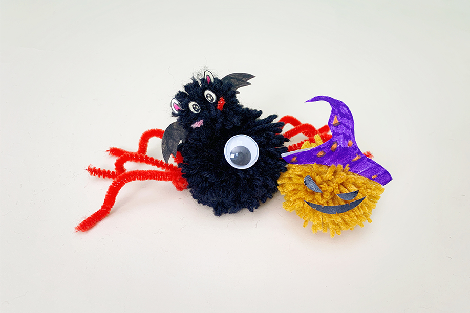 Two completed Halloween-themed pom-pom creatures are presented on a white surface. One is a black pom-pom spider with red pipe cleaner legs and a small bat attached to its body, complete with paper wings and features. The other is a yellow pom-pom jack-o'-lantern wearing a purple paper witch's hat. Both are charmingly crafted, showcasing simple yet effective Halloween-themed designs. The overall impression is one of fun, handmade decorations.