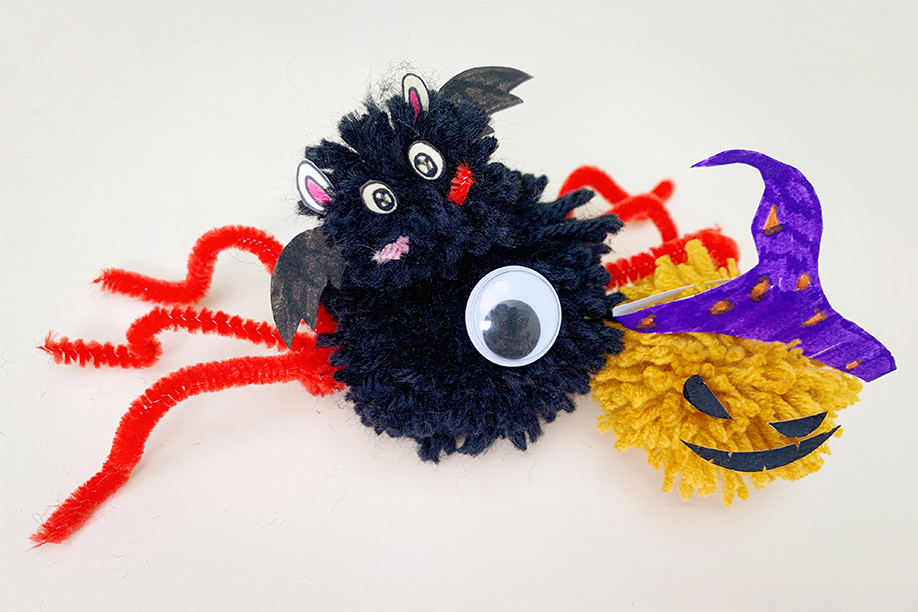 Two charming Halloween-themed pom-poms are artfully arranged on a white surface. A black pom-pom spider, with bright red pipe cleaner legs, features a smaller black bat pom-pom affixed to its body; the bat has paper wings and facial features. Beside it sits a yellow pom-pom jack-o'-lantern, sporting a purple paper witch's hat. Both are meticulously crafted, demonstrating simple yet effective Halloween designs. The overall impression is one of delightful, handmade decorations, perfect for a festive display.