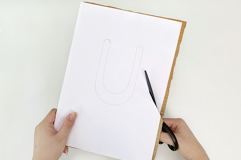 Two hands carefully use a pair of black scissors to cut out a drawn shape from a piece of white paper. The shape is a large, rounded 'U', lightly sketched onto the paper. The paper is supported by a piece of cardboard, and the hands hold the paper steady while cutting along the pencil line. The scene is set against a plain white background, drawing attention to the precise cutting action. The overall impression is one of a calm, controlled step in a craft project.