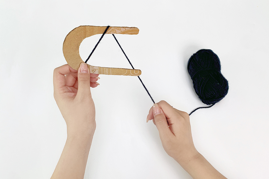 Two hands are shown wrapping dark blue yarn around a U-shaped piece of cardboard. One hand holds the cardboard steady whilst the other hand carefully winds the yarn around the frame, creating a pom-pom. A ball of the same yarn sits to the side. The background is plain white, drawing focus to the hands and the process of pom-pom creation. The image depicts a clear step in a craft project.