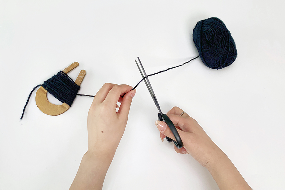 Two hands are shown using scissors to cut a length of dark blue yarn. A U-shaped cardboard pom-pom maker, already wound with the same yarn, is visible to the left, and a ball of the yarn rests to the right. The hands hold the yarn taut as the scissors precisely sever it. The background is a plain white surface, focusing attention on the cutting action. The image depicts a step in the process of making a yarn pom-pom.