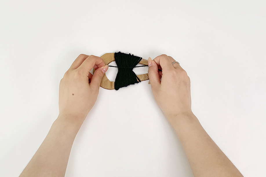 Two hands are carefully tying off a bundle of dark blue yarn that has been wound around a U-shaped cardboard frame. The yarn is neatly gathered in the centre of the frame, and the hands are gently securing it with a knot. The background is a plain white surface, which makes the hands and the yarn the central focus. The image shows a precise step in the process of making a yarn pom-pom.