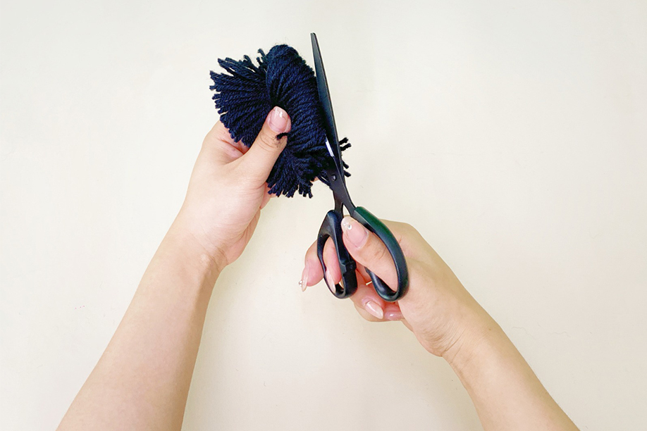 Two hands are shown trimming a dark blue yarn pom-pom. One hand holds the pom-pom, while the other hand uses small, sharp scissors to carefully cut the loose yarn ends, creating a more even and defined shape. The background is a plain, off-white surface, which draws attention to the precise cutting action. The image depicts a detail-oriented step in the pom-pom making process, focusing on achieving a neat and polished finish.