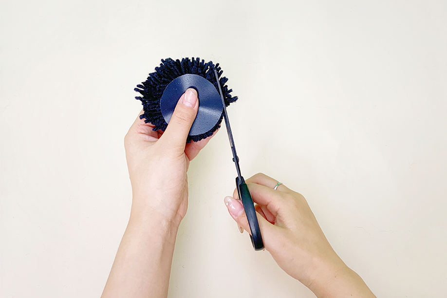 Two hands carefully use scissors to trim the excess yarn from a dark blue yarn pom-pom. The pom-pom is held firmly in one hand, while the other hand uses the scissors to create an even, neat edge around the circular shape. A small, dark disc is visible in the centre of the pom-pom, suggesting the use of a cardboard template. The background is a plain white surface, highlighting the precise cutting action. The image shows a meticulous step in the pom-pom making process.