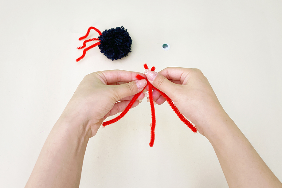 Two hands are shown manipulating two red pipe cleaners against a plain white background. A dark blue yarn pom-pom and a single googly eye are visible in the background. The hands are carefully twisting the pipe cleaners together near their centres, preparing to attach them to the pom-pom to create spider legs. The image focuses on this precise step in the craft project's construction.