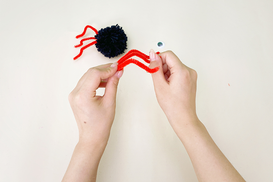 Two hands are gently bending a single red pipe cleaner into a curved shape. A dark blue yarn pom-pom and a single googly eye are visible in the background, suggesting the creation of a spider. The hands are carefully manipulating the pipe cleaner to achieve the desired curve, preparing it to be attached as a leg to the pom-pom. The plain off-white background focuses attention on the hands and the pipe cleaner. The image shows a step in the construction of a simple craft project.