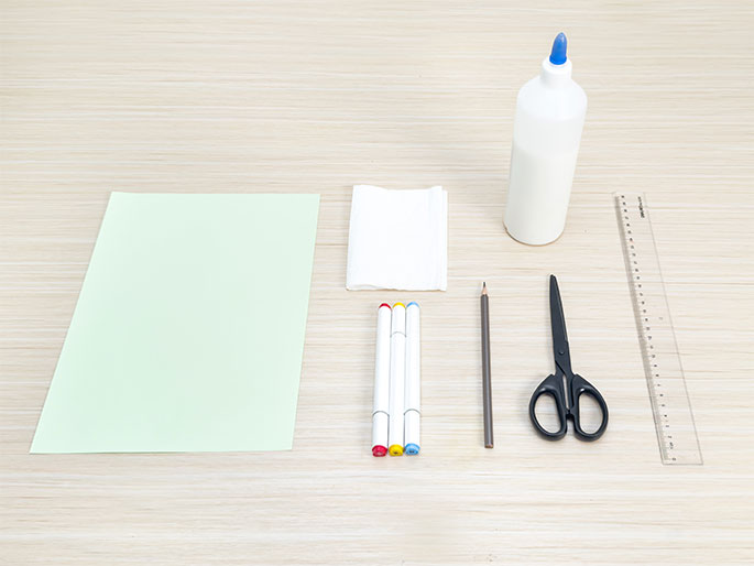 An assortment of craft supplies arranged on a light wooden table. The supplies include a pale green sheet of paper, a folded white tissue paper, a bottle of white glue, a clear plastic ruler, a pair of black scissors, a pencil, and three white markers with red, yellow, and blue caps. These items suggest preparation for a paper craft project.