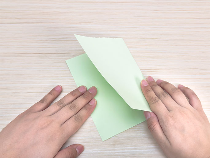 Two hands are folding a pale green piece of paper in half on a light wood table. This image shows the first step in a paper folding activity.