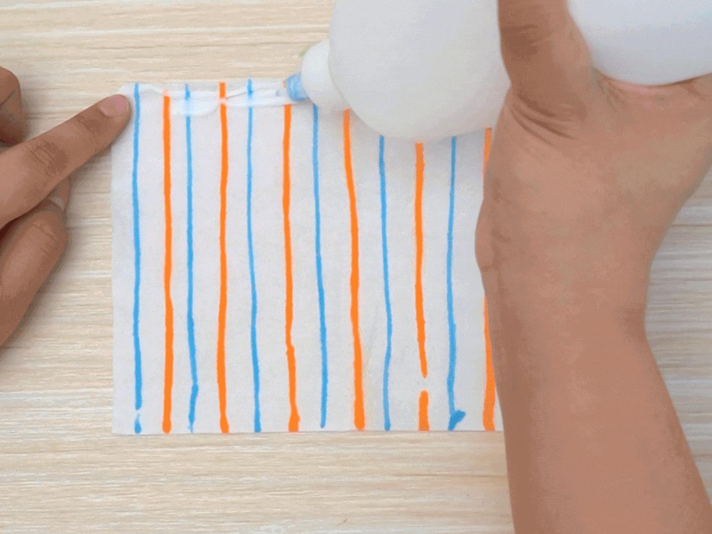 Glue is being applied along the top edge of a white rectangular piece of paper decorated with alternating orange and blue horizontal stripes. One hand holds the glue bottle while a finger of the other hand keeps the paper in place on a light wood grain surface. This image shows a step in a crafting process, likely involving gluing the paper to another object.