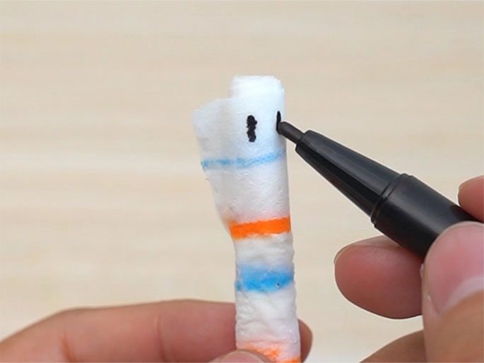 A black marker held in one hand draws two small vertical lines on the top edge of a paper-covered stick held between the fingers of another hand. The paper has horizontal stripes in orange and blue. This close-up image depicts the process of adding details, likely eyes, to a handmade craft figure.