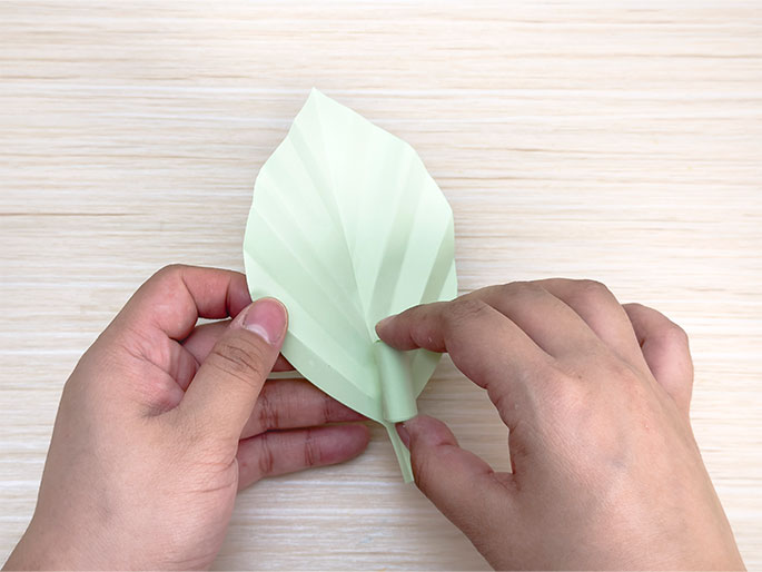 Two hands shape a pale green origami leaf on a light wood grain surface. The fingers pinch the base of the leaf where it narrows into a stem-like shape. This image captures the delicate process of folding and shaping paper in the art of origami.