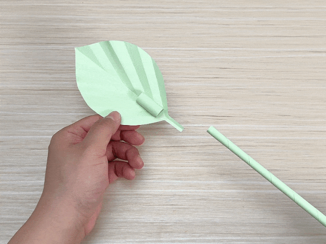 A hand holds a pale green origami leaf next to a pale green paper-covered stem on a light wood grain surface. The base of the leaf has a small opening where it appears designed to attach to the stem. This image shows the components of a paper flower or plant craft before assembly.