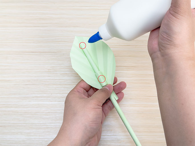 Glue is being applied from a white bottle with a blue tip to the base of a pale green origami leaf. One hand holds the leaf and stem while the other applies the glue, preparing to attach the leaf to the stem. This image shows a step in assembling a paper flower or plant craft on a light wood grain surface.