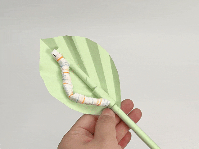 A hand holds a completed craft project featuring a pale green origami leaf attached to a pale green stem. A small, striped paper caterpillar, made with orange, blue, and white stripes, rests on the leaf. The background is a solid light gray or white. This image showcases a finished paper craft, likely depicting a caterpillar on a leaf.