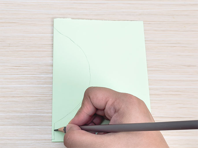 A hand draws a curved line with a pencil on a light green piece of paper folded in half. The paper rests on a light wood table. This image shows the process of drawing a shape on folded paper, likely to create a symmetrical design when unfolded.