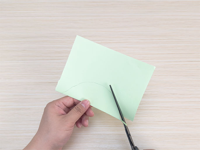 A hand holds a piece of folded light green paper while another hand cuts along a curved line drawn on the paper with scissors. The paper is positioned on a light wood table. This image demonstrates cutting a folded piece of paper to create a specific shape.