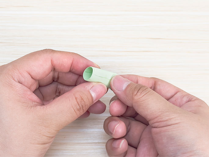 Two hands hold a small, pale green cylinder made of rolled paper. The cylinder is held horizontally above a light wood grain surface. This image shows the next stage in a paper craft project.