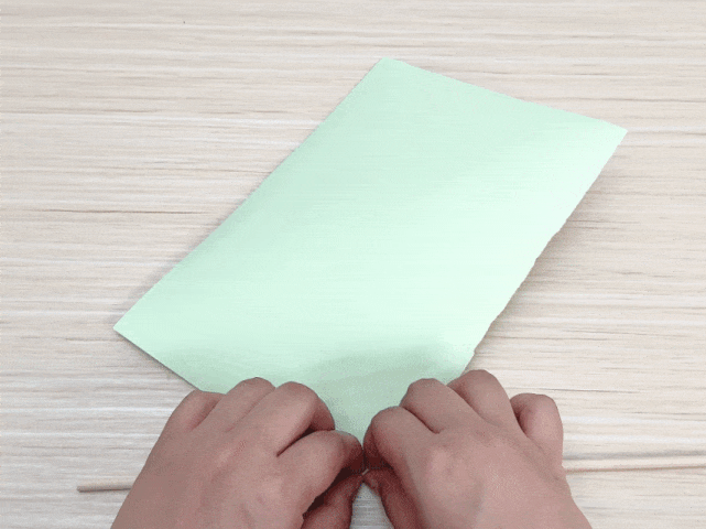 Two hands are folding a piece of pale green paper in half lengthwise on a light-colored wooden table. A small section of a thin wooden dowel is visible at the bottom edge of the paper. This image captures a step in a paper folding or craft activity.