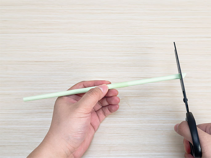A pair of scissors held in one hand cuts the end of a long, thin, pale green tube held in the other hand. The tube and scissors are positioned above a light wood grain surface. This image demonstrates the process of trimming or cutting a paper tube for a craft or other project.