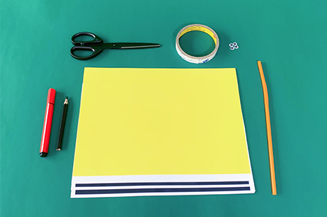 Arranged on a green surface are various craft supplies, suggesting preparation for a DIY project. A large sheet of yellow paper, accented with two dark blue stripes at the bottom, takes center stage. Beside it lie a pair of black scissors, a roll of double-sided tape, a red marker, a pencil, a few small metal brads, and a long, thin orange stick or dowel. This flat lay presentation clearly displays the materials needed for the upcoming activity.