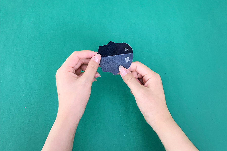 In this image, a person is holding two pieces of black paper that have been cut into the shape of a bat's body and wings. Small pieces of double-sided tape are visible, ready to be used to attach the parts together. This simple paper bat craft is ideal for Halloween, allowing crafters of all ages to create their own spooky decorations with minimal materials, such as paper, scissors, and tape. The finished bat can be used to decorate walls, windows, or other areas, adding a creative and festive touch to any Halloween setting.