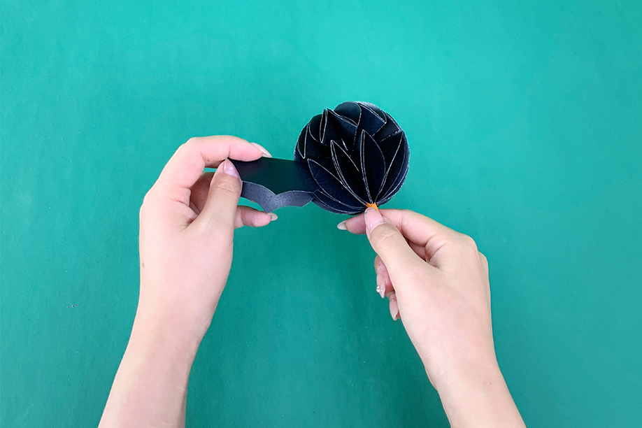 In this image, a person is attaching the wings to a 3D paper bat, which has been crafted by folding black paper into a spherical shape for the body. The wings are precisely cut and being carefully positioned to complete the bat's structure. This simple and creative Halloween craft is perfect for both children and adults, requiring only basic materials such as paper, scissors, and glue. Once finished, the paper bat can be used as a decoration, adding a spooky and festive touch to any Halloween celebration or display.