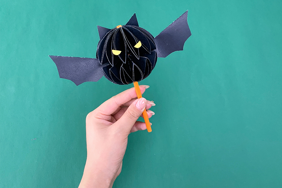 In this image, a person is holding a completed 3D paper bat craft attached to an orange straw. The bat's body is made from folded black paper, giving it a spherical, layered appearance, while its wings and pointed ears add to the spooky design. Yellow eyes have been added to give the bat a playful yet eerie look, perfect for Halloween decorations. This craft is simple to make and ideal for children and adults alike, requiring minimal materials such as paper, scissors, glue, and a straw. The finished bat can be used as a fun decoration or prop for Halloween festivities.