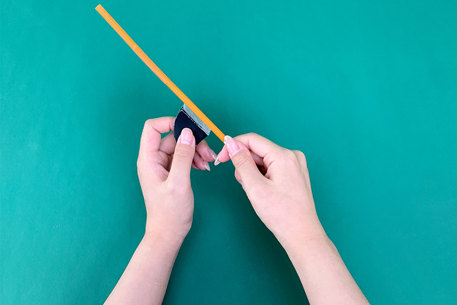 In this image, a person is attaching a small paper bat to a pencil using a piece of double-sided tape, creating a fun and interactive craft. The bat, made from black paper, will flutter when the pencil is moved, making it a playful addition to any Halloween celebration. This simple yet creative craft is perfect for children, allowing them to combine art and practical objects like pencils. With minimal materials required—just paper, tape, and a pencil—it’s an easy and enjoyable way to add a spooky touch to everyday items during the Halloween season.