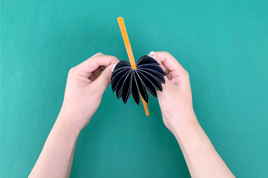 In this image, a person is holding a completed paper bat craft, which has been attached to a pencil. The bat is made from layered black paper, folded and fanned out to create a 3D effect, with the pencil serving as a handle or central support. This fun and easy project is ideal for Halloween, allowing crafters to make their own decorative bats with minimal materials. The finished bat can be used as a playful prop or decoration, adding a spooky yet charming touch to any Halloween setting. It's a perfect activity for children and adults alike, encouraging creativity and hands-on crafting.