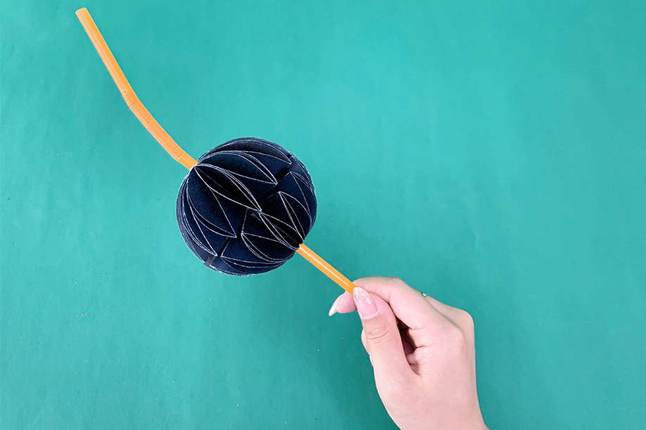 In this image, a person is holding a completed 3D paper bat craft attached to a pencil. The bat is formed by folding and layering black paper into a spherical shape, creating a striking visual effect. This simple yet creative craft is perfect for Halloween, offering an engaging activity for children and adults alike. With just a few materials, such as paper, glue, and a pencil, this DIY project adds a playful and spooky touch to everyday items. Ideal for classroom activities or home decorations, this paper bat craft brings a festive Halloween vibe to any setting.