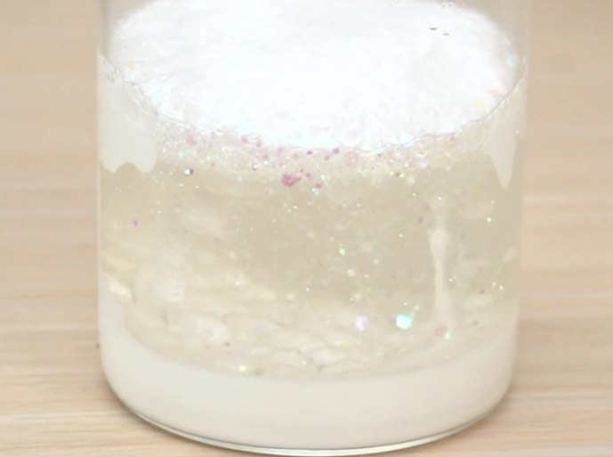 A clear glass jar sits on a light wooden table. The jar contains a layered mixture: a white, opaque substance at the bottom, a clear liquid layer in the middle, and a thicker, white substance at the top. Some of the top layer drips down into the clear liquid, creating a visually interesting effect. Small, sparkling particles are visible within the clear liquid layer. The video showcases the mixing or layering of different substances, possibly a science experiment or the creation of a sensory toy.