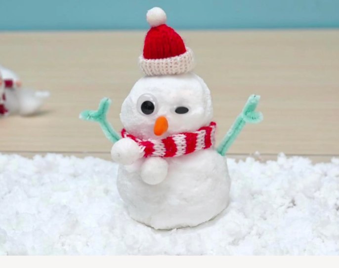 A charming snowman sits on a bed of fluffy white snow against a light blue backdrop. The snowman is adorned with a red and white knitted hat and scarf, googly eyes, an orange carrot nose, light green twig arms, and small white snowball hands. A miniature Santa Claus figure is faintly visible in the background, slightly out of focus. A wooden surface is also visible beneath the snow.