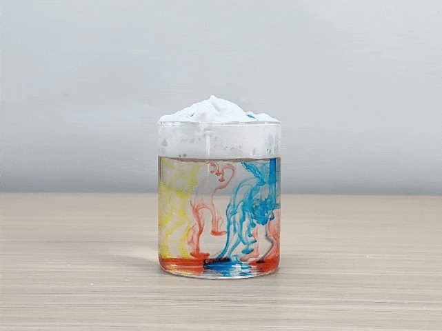 A clear glass, partially filled with water, sits on a light-colored wooden surface. A layer of white powder, possibly baking soda or another similar substance, rests on top of the water. A hand carefully drops blue food coloring onto the white powder. The color slowly seeps down through the powder and creates swirling patterns in the water below. The video appears to be demonstrating a simple science experiment, possibly illustrating the principles of density or diffusion.