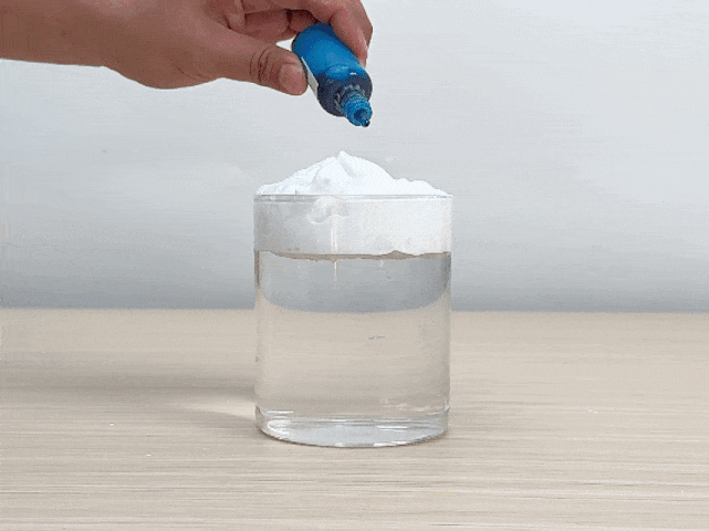 A clear glass, partially filled with water, sits on a light-colored wooden surface. A layer of white powder, possibly baking soda or another similar substance, rests on top of the water. A hand carefully drops blue food coloring onto the white powder. The color slowly seeps down through the powder and creates swirling patterns in the water below. The video appears to be demonstrating a simple science experiment, possibly illustrating the principles of density or diffusion.
