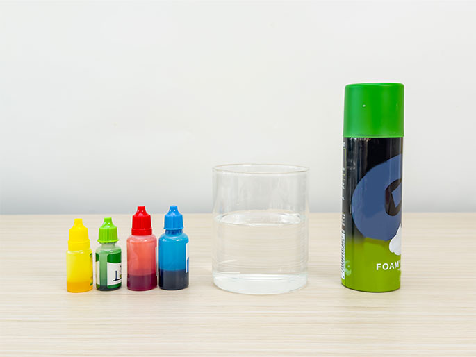 Arranged on a light wooden table are several items, suggesting preparation for a science experiment or craft activity. Four small bottles containing colored liquids—yellow, green, red, and blue—are lined up. Next to them is a clear glass half-filled with water. A green can, likely containing shaving cream or a similar foaming substance, sits beside the glass. The scene implies an upcoming demonstration involving color mixing or a creative project using these materials.