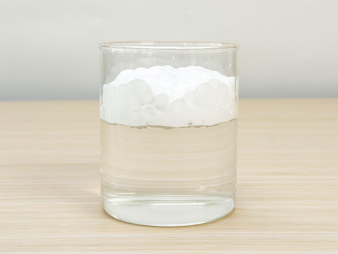 A clear glass sits on a light wood grain table. The glass is partially filled with water, and a thick layer of white foam sits on top, resembling a cloud or a mound of whipped cream. This simple setup suggests a science experiment demonstrating density or a visual representation of layers.