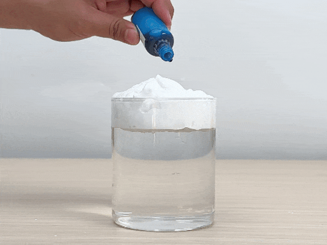A clear glass, partially filled with water, sits on a light wooden surface. A layer of white powder, resembling baking soda or a similar substance, floats on top of the water. A hand gently squeezes drops of blue food coloring onto the white powder. The blue coloring slowly permeates the powder and begins to seep into the water below, creating mesmerizing swirls and patterns. This video appears to demonstrate a simple science experiment, possibly illustrating the principles of density, diffusion, or solubility.