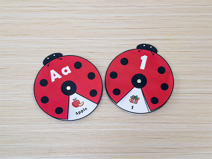 Two completed ladybug-shaped learning spinners are displayed on a light wood-grain table. The left spinner shows the letters Aa and a picture of an apple slice with the word Apple. The right spinner shows the number 1 and a picture of a gift with the number 1. These spinners are designed as educational tools for young children, likely for matching or alphabet and number recognition activities.