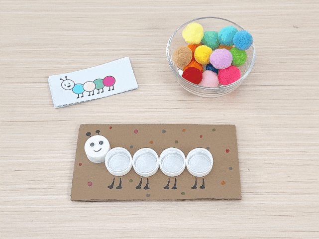 A hand uses blue plastic tongs to place colorful pom-poms into white bottle caps glued to a piece of brown cardboard. The bottle caps and pom-poms form a caterpillar, with the first bottle cap having a drawn-on face. A small bowl of colorful pom-poms sits beside the caterpillar, and a drawing of a similar caterpillar is visible in the corner. This video demonstrates a simple children's activity promoting fine motor skills and color recognition.