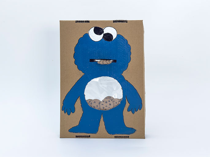 Against a plain white background stands a finished Cookie Monster figure made from cardboard. Painted blue, the Cookie Monster has a slit for a mouth, where a small portion of a chocolate chip cookie is visible. The figure's belly has a circular window covered with clear plastic, revealing more chocolate chip cookies inside. This appears to be a completed cardboard Cookie Monster feeding toy or game.
