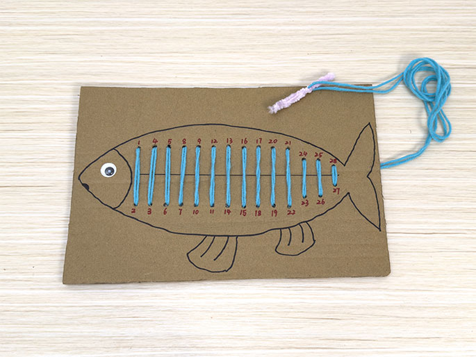 A fish-shaped piece of brown cardboard rests on a light wood-grain table. The fish has a googly eye and black outlined fins. Numbered holes are punched around the fish's outline, and teal yarn has been laced through most of the holes, creating vertical stitches across the body. A small section at the top right, and the bottom edge remain unlaced. A light purple pipe cleaner is tied to the end of the yarn at the top. This image depicts a partially completed children's sewing or lacing activity.