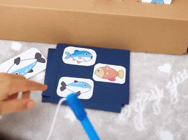 A child plays a fishing game. The game board, made from a cardboard box, has slots containing colorful fish cutouts, each with a small paperclip attached. A child's hand uses a toy fishing rod, made from a blue stick and string with a magnet at the end, to catch the fish. The video also shows the child interacting with separate fish cards, likely used for matching or identification before or during gameplay. The game is set on a light-colored surface with a subtle heart pattern. The video demonstrates a simple and engaging activity, likely designed for young children, promoting hand-eye coordination and color recognition.