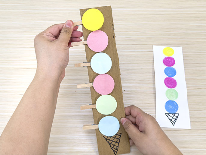 Two hands interact with a cardboard ice cream cone craft on a light wooden surface. The cardboard has a hand-drawn ice cream cone at the bottom and several small wooden clothespins clipped along its length. Each clothespin holds a colorful paper circle, representing a scoop of ice cream: yellow, pink, light blue, pink, light green, and light blue from top to bottom. To the right, a separate piece of white paper shows a similar ice cream cone design with painted circles in yellow, pink, blue, pink, green, and blue, likely serving as a model for the cardboard version. This image captures a crafting activity, possibly a learning game for color recognition or sequencing.