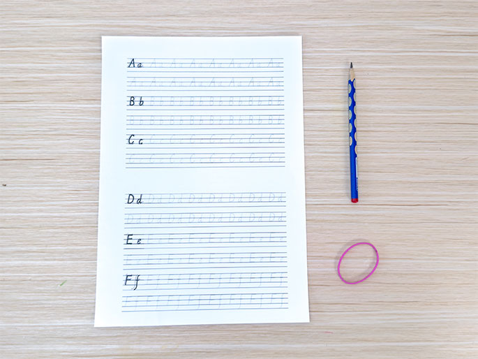 A sheet of paper with handwriting practice lines lies on a light wood-grain table. The paper displays rows of cursive letters from A to F, both uppercase and lowercase, with dotted outlines for tracing. Beside the paper, a blue pencil with a patterned design and a pink rubber band are also on the table. This image suggests a setting for handwriting practice or a lesson in cursive writing.