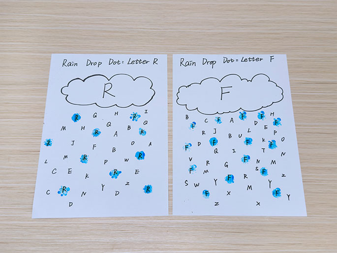 Two sheets of white paper, titled Rain Drop Dot: Letter R and Rain Drop Dot: Letter F, lie on a light wood-grain surface. Each sheet features a hand-drawn cloud at the top, containing the respective letter (R or F). Below each cloud, grids of letters are presented. All instances of R on the first sheet and F on the second sheet are marked with blue paint dots. This image depicts a completed letter recognition activity or game for children.
