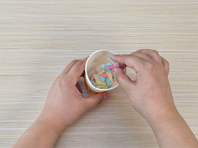 Two hands interact with a small white paper cup containing small, colorful pieces of cut paper on a light wood-grain surface. The cup is partially wrapped with orange tape near the bottom. One hand holds the cup while the other picks up some of the colorful paper pieces, suggesting they are being added or arranged. The colors of the paper pieces are pastel pink, blue, and yellow. This image likely depicts a step in a craft activity.