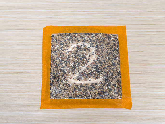 A clear plastic bag filled with mixed rice grains and edged with orange tape sits on a light wood-grain surface. The number 2 is clearly traced into the rice, revealing the lighter surface below. This image likely demonstrates a sensory activity or learning tool using a rice-filled bag for tracing letters or numbers.