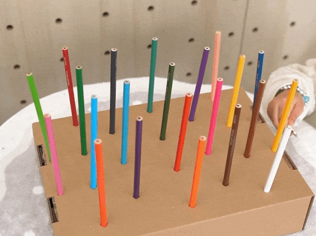 A small child's hand interacts with a homemade game. The game consists of a cardboard box with numerous holes, each containing an upright colored pencil. The child's hand places a coin between the pencils. The pencils vary in color, creating a vibrant and engaging visual. The game likely involves dropping the coin and seeing where it lands, perhaps associating a color or point value with each pencil. The background is a blurred light wooden surface.