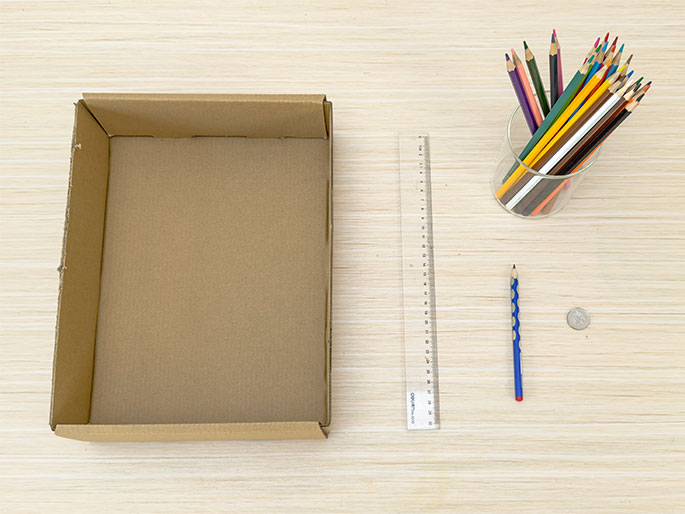 A flat, open cardboard box lies on a light wooden table. Next to it are a clear plastic ruler, a blue pencil, and a small silver coin. A clear glass or jar containing a collection of colored pencils sits near the top right corner. The arrangement suggests the preparation for a craft or construction project.